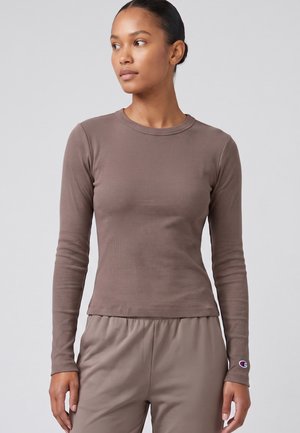 Frau mit Dutt trägt ein eng anliegendes, langärmeliges, geripptes Shirt in Taupe und passende Hosen, sie schaut leicht nach links vor einem grauen Hintergrund.