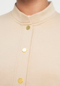 Ανώτερο μέρος σε κρεμ απόχρωση με όρθιο γιακά, χρυσά κουμπιά με ομαλή υφή και ραφές κατά μήκος της μπροστινής πατιλέτας.