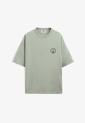 Personne aux cheveux courts, vue de dos, portant un t-shirt oversize vert clair avec le texte « product of berlin » dans le dos.