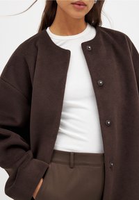 Braune Fleece-Jacke mit rundem Ausschnitt und Druckknopfverschluss, getragen über einem weiß gerippten Oberteil, kombiniert mit braunen Hosen.