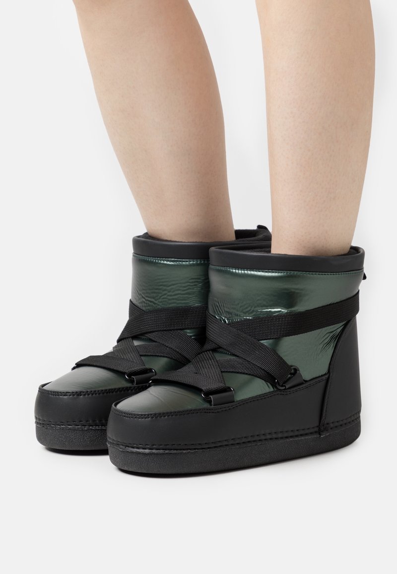 Jambes portant des bottines rembourrées brillantes vert foncé et noires avec de larges sangles croisées noires sur un fond blanc.