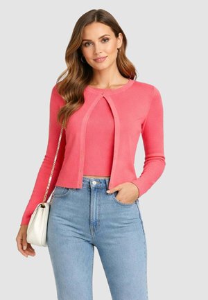 Donna che indossa un cardigan rosa corto sopra un top abbinato, jeans azzurri a vita alta, tiene una borsa bianca con tracolla a catena.