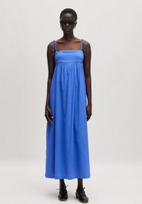 Blaues Maxikleid mit verstellbaren Trägern, strukturiertem Stoff und gerafftem Taillengummi, kombiniert mit schwarzen Schnürschuhen.