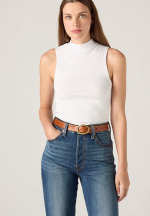 Femme portant un haut blanc sans manches à col montant, un jean bleu et une ceinture en cuir marron avec une boucle dorée, debout avec une main dans la poche.