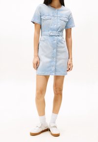 Modré pruhované denimové šaty s krátkými rukávy, zapínáním na knoflíky vpředu, pasem s páskem a dvěma náprsními kapsami. Kombinované s bílými teniskami a ponožkami.