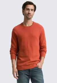 Mann steht mit einer Hand in der Tasche, trägt einen orangefarbenen Langarmpullover und blaue Jeans vor einem schlichten hellen Hintergrund.