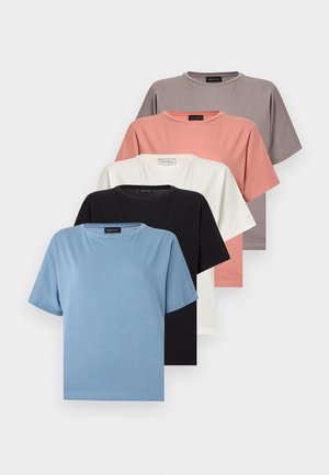 Γυναίκα με γκρι κοντομάνικη μπλούζα, μπλε τζιν με φαρδιά πόδια, μαύρα παπούτσια, χρυσούς κρίκους σκουλαρίκια και μαύρα ορθογώνια γυαλιά ηλίου, στέκεται με τα χέρια πίσω από την πλάτη.