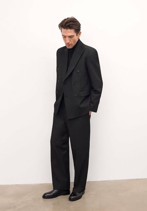 Jachetă de costum neagră, cu două rânduri de nasturi, și pantaloni ajustați, confecționată dintr-un material neted. Include un top negru cu guler înalt dedesubt și pantofi din piele.
