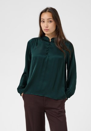 Femme portant un chemisier vert foncé à manches longues avec des boutons et un pantalon marron, debout devant un fond clair uni.