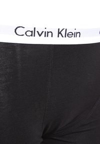 Crne tajice od pamučne mješavine s rebrastom teksturom i bijelim pojasom na kojem je u crnoj boji napisano "Calvin Klein". Bez dodatnih uzoraka ili naglasaka.