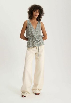 Femme aux cheveux bouclés portant un haut sans manches à motifs avec un col en V et un pantalon large crème, debout les mains dans le dos.