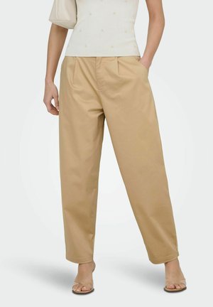 Vrouw met losvallende beige broek, wit mouwloos topje, sandalen met doorzichtige bandjes en een lichtbeige schoudertas op een witte achtergrond.