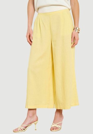 Kobieta ubrana w luźne, z wysokim stanem, szerokie żółte spodnie w zestawieniu z żółtymi sandałami na obcasie z paskami i białym topem.