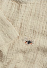Beige tyg med en strukturerad yta som har en liten, broderad koala i ljusrosa och svart, med röda accenter på kinderna.