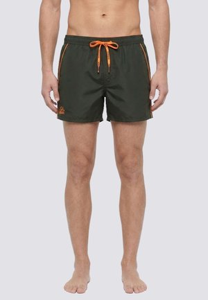 Uomo con pantaloncini da bagno verde scuro con rifiniture arancioni e coulisse, in piedi a piedi nudi su uno sfondo semplice.