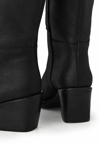 Botas negras hasta la rodilla, fabricadas en material sintético texturizado, con un tacón grueso de diseño acanalado y detalles de costura sutiles.