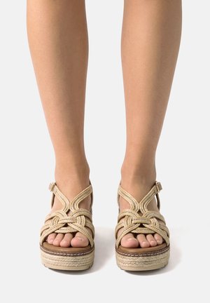 Pies con sandalias beige de plataforma trenzada con correas en el tobillo, sobre un fondo blanco liso.