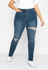 Jeans skinny de color azul oscuro, de cintura alta, con desgastes y rotos en las rodillas, y bolsillos laterales. Llevados con zapatillas de lona blancas. Material de mezcla de algodón.