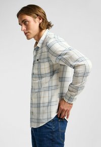 Ljust rutigt skjorta i krämfärg med blå och gula linjer, med långa ärmar, knäppning i kragen och en bröstficka.