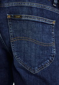 Mørkeblå denimjeans med en karakteristisk lomme, kontrasterende gul søm og en liten svart "Lee" etikett på øvre kant.