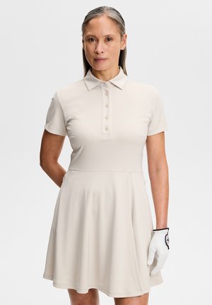 Kvinde iført en beige polokjole med korte ærmer og en hvid golfhandske på venstre hånd mod en ensfarvet baggrund.