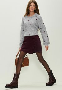 Šedý sveter s motívom srdca, bordová sukňa s viazaním, čierne nadkolienkové čižmy a hnedá kabelka. Hladká textúra, ležérny štýl.