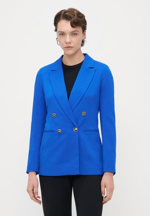 Blue double-breasted blazer in a smooth fabric, featuring gold buttons and notch lapels, paired with a black top and black trousers.