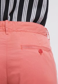 Różowe spodnie z bawełny o tapered designie, z tylną kieszenią zapinaną na guzik i paseczkami, wyeksponowane na tle czarno-białej koszuli w paski.