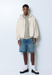 Beige Kapuzenpullover über einem grün karierten Hemd, kombiniert mit Denim-Shorts und schwarzen Loafern mit weißen Sohlen und Socken.