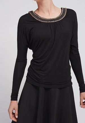 Femme portant un haut noir à manches longues avec un col décoratif en chaîne dorée et noire et une jupe noire.