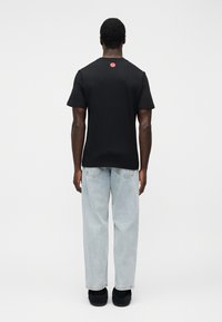 Μαύρο βαμβακερό t-shirt  με κοντά μανίκια, με ένα κόκκινο κυκλικό λογότυπο στην πλάτη, συνδυασμένο με ανοιχτό μπλε, χαλαρά τζιν.