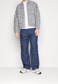 Herre quiltet jakke med et marineblåt og hvidt mønster, kombineret med løse blå bukser og hvide sportssko med sorte detaljer.