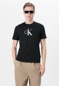 Zwarte katoenen t-shirt met een zilveren "CK" logo. Korte mouwen, klassieke pasvorm, gedragen met beige broeken. Model draagt zwarte zonnebril.