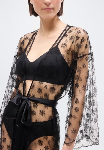Γυναίκα που φοράει μαύρο floral δαντελένιο ρόμπα δεμένο με σατέν κορδέλα πάνω από αντίστοιχο μαύρο σουτιέν και εσώρουχο, σε απλό φόντο.