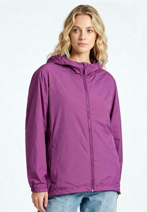 Giovane donna con capelli biondi mossi che indossa una giacca viola con cappuccio e cerniera e jeans azzurri, che guarda leggermente verso destra su uno sfondo semplice.