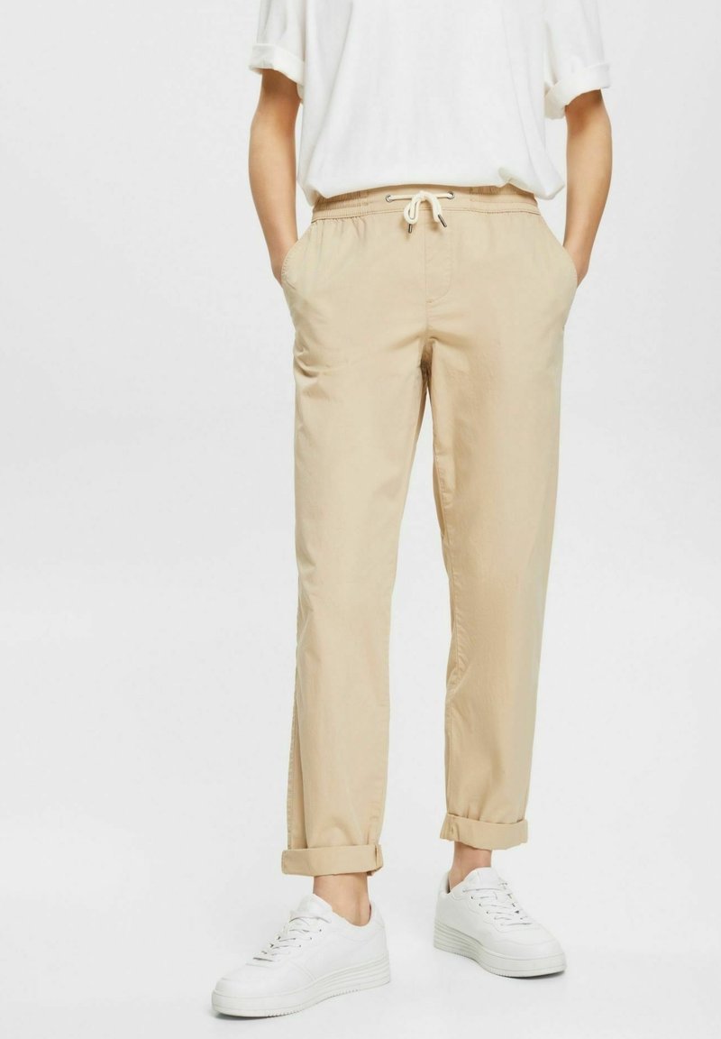 Personne portant un pantalon beige à cordon de serrage roulé aux chevilles, une chemise blanche à manches courtes et des baskets blanches, debout avec les mains dans les poches.
