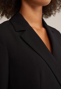 Svart kavaj med notchrev, strukturerade axlar och en mjuk textur, som visar en djup V-ringning och skräddarsydd passform.