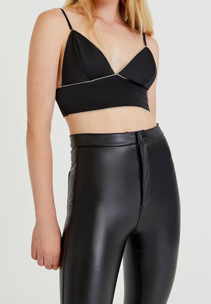 Top corto negro con tirantes finos y adornos de pedrería. Combinado con leggings de cuero sintético negro brillante de tiro alto con cremallera frontal.