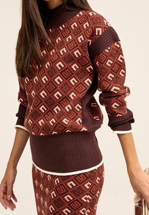 Femme portant un pull et une jupe assortis à motif géométrique marron et rouille avec des poignets et un ourlet côtelés, tenant un sac beige dans une main.