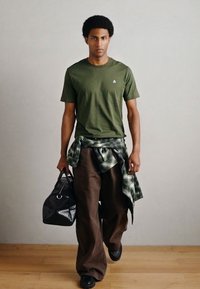 Πράσινο βαμβακερό T-shirt, καφέ φαρδιά παντελόνια, μαύρη τσάντα με υφή, καρό πουκάμισο δεμένο στη μέση και μαύρα παπούτσια. Απλό, καθημερινό ντύσιμο.