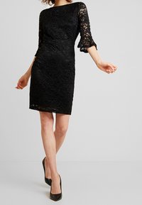 Svart spetsklänning med trekvartslånga ärmar, figurnära midja och utsvängda ärmslut. Spetsmönstret har blommönster. Längd ovanför knäna. Svarta klackar.