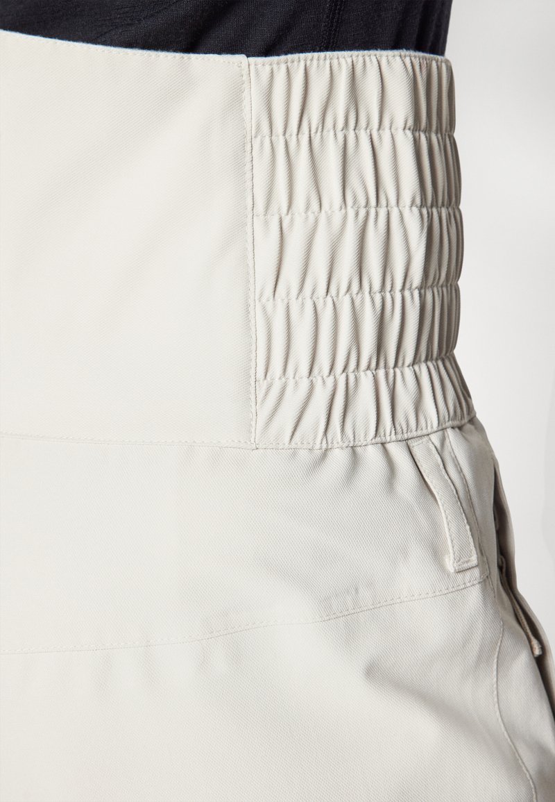 Jasnobeżowe spodnie z szerokim, marszczonym paskiem w talii. Materiał wydaje się teksturowany, o gładkim wykończeniu, z boczną kieszenią.