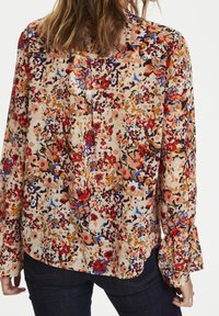 Mujer vestida con una blusa floral de mangas largas en tonos rojos, naranjas, azules y beiges, combinada con jeans de color azul oscuro, mostrada desde la espalda.