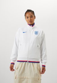 Hvit zip-up jakke med blå Nike-logo og England-skjold. Har røde og blå detaljer på kragen og mansjettene. Myk, glatt stoff.