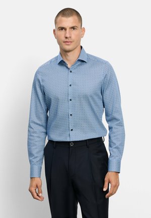 Man met een aansluitend blauw patroond hemd met lange mouwen en zwarte broek, staand tegen een effen lichte achtergrond.