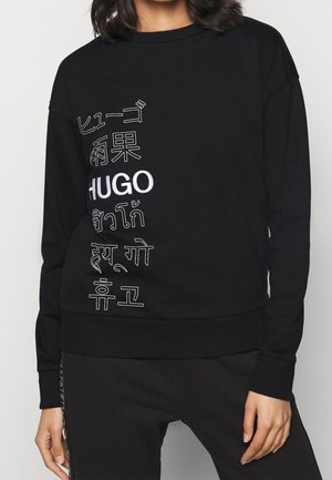Zwarte lange mouwen sweat met "HUGO" en meertalige tekst verticaal gedrukt op de voorkant, gedragen door een persoon met losse zwarte broek.