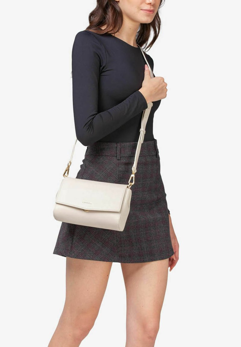 Borsa a tracolla in pelle beige con chiusura a pattina, una fibbia dorata e una lunga tracolla regolabile, indossata da una persona che veste un top nero e una gonna a quadretti.