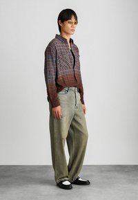 Camicia di flanella a quadri in tonalità rosse e blu, jeans oversized di un verde chiaro e scarpe nere e bianche. Gli accessori includono occhiali da sole colorati.