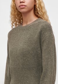Jeune personne aux longs cheveux blonds bouclés portant un pull en maille vert olive uni sur fond blanc.