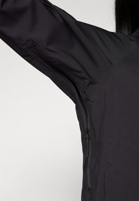 Black jacket featuring a textured fabric, zippered arm pocket, and sleek silhouette, designed for functionality and comfort.